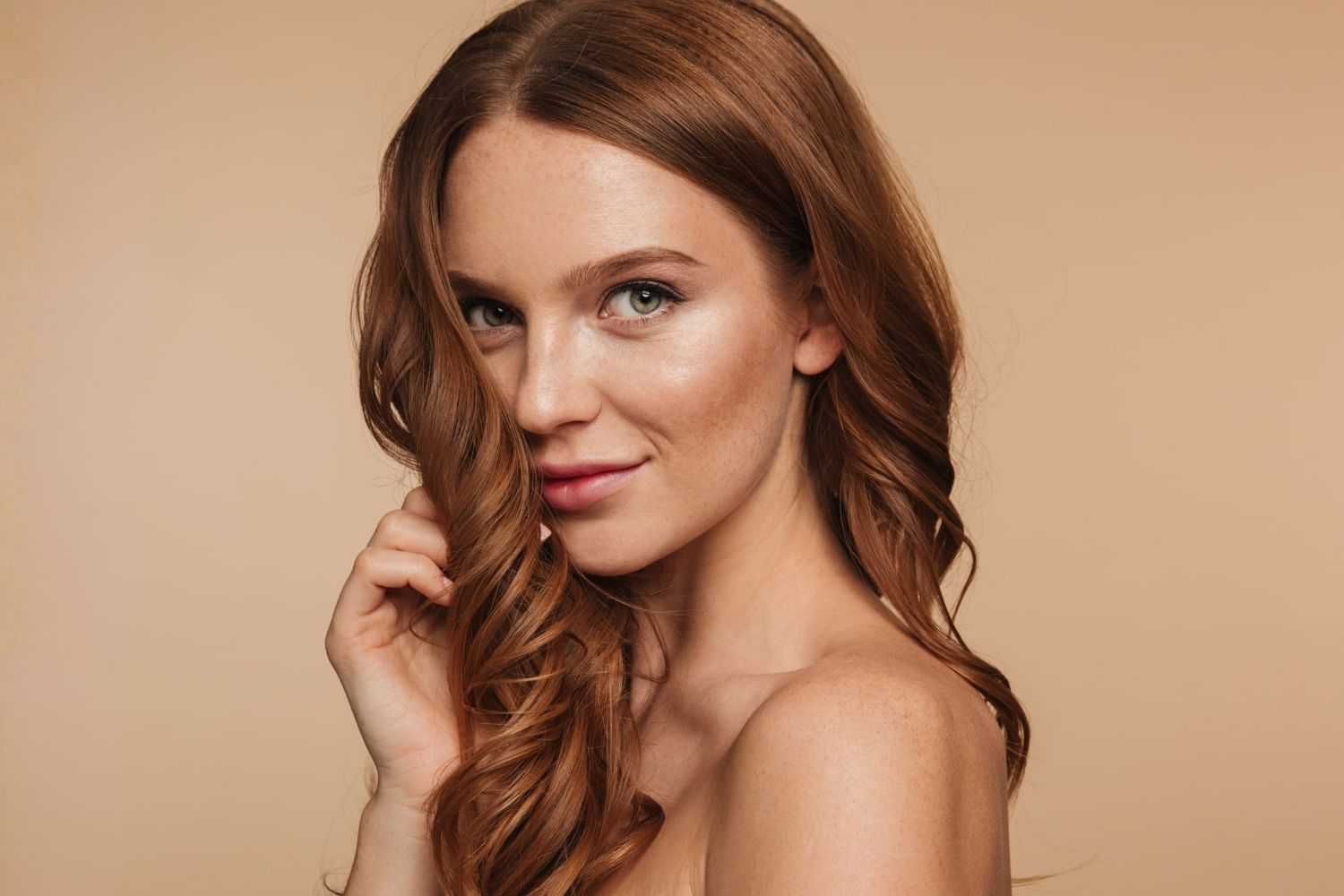Young woman with long red hair smiling in front of a beige background.
