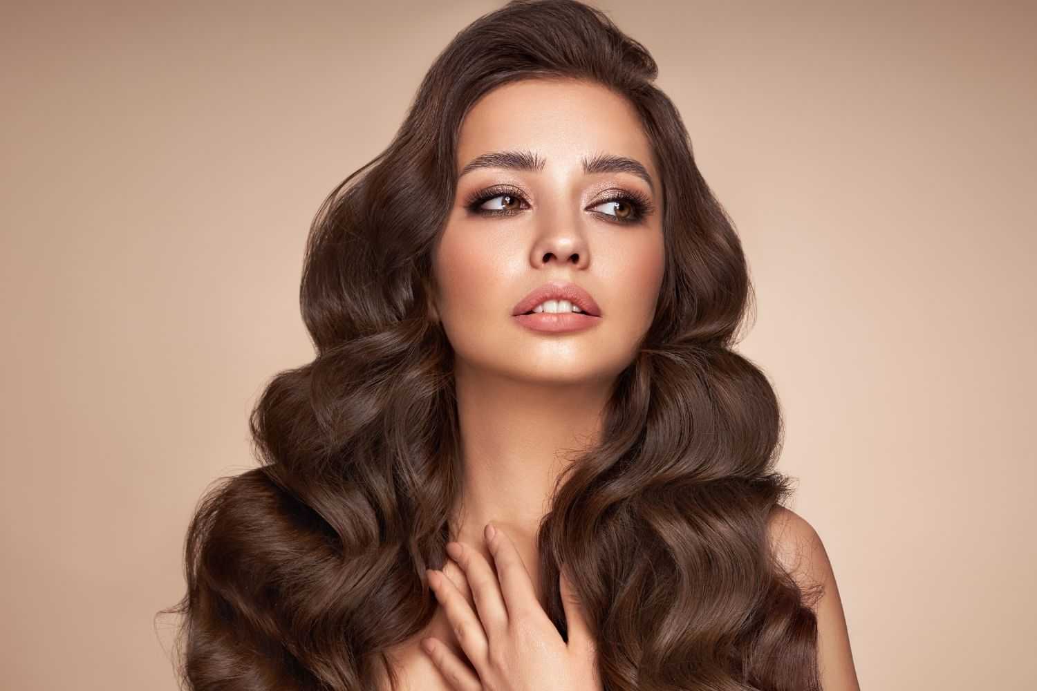 Woman with long, wavy brown hair and neutral makeup on a beige background.