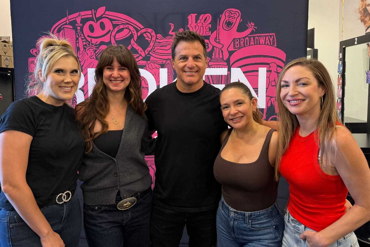 Group of five people smiling in front of a pink and black backdrop with graphics and text.